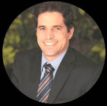 Smiling man with dark wavy hair in a black suit and striped tie, blurred greenery.