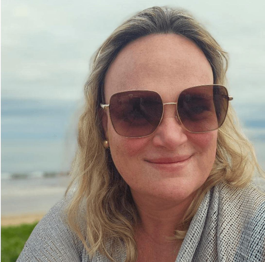 Blonde woman in large square sunglasses and grey mesh top smiling with beach background.